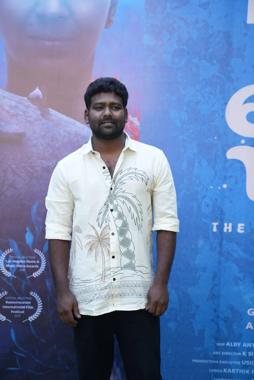 An Enna Vilai cast member in a white shirt stands before the film's trailer artwork