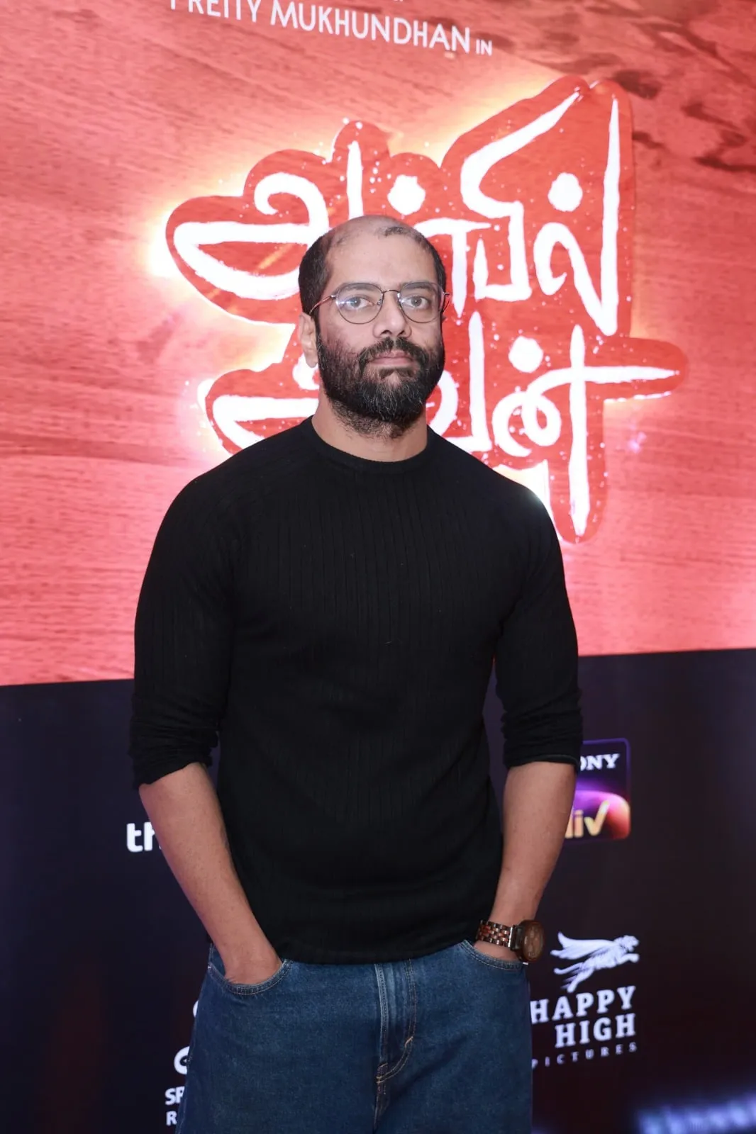 Portrait with glasses and a black pullover against the Anbil Avan backdrop