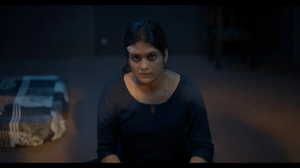 A young woman in a dark kurta with a head bandage, sitting in a dim interrogation-style room.