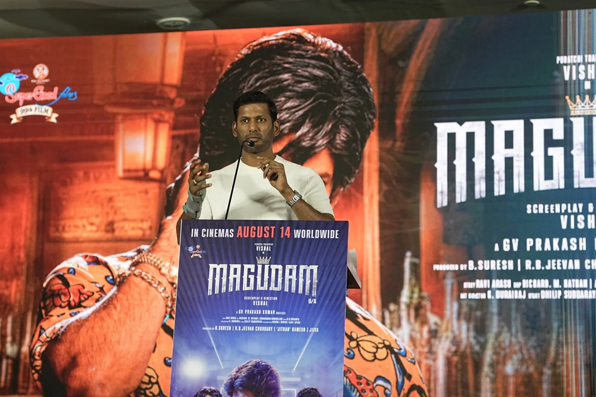 Vishal speaking at the Magudam trailer launch podium, with his aged look from the film on the screen behind him